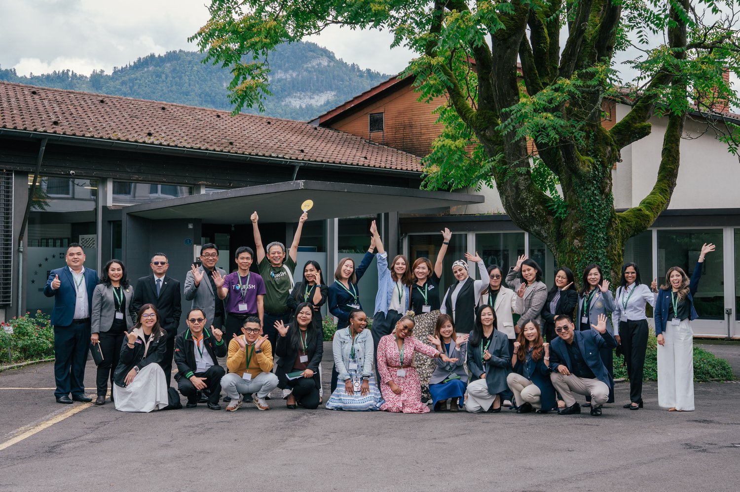 Group photo of participants at the IMI Switzerland Partners & Counselors Event 2025, standing and crouching outdoors in front of a building with a tree and mountains in the background